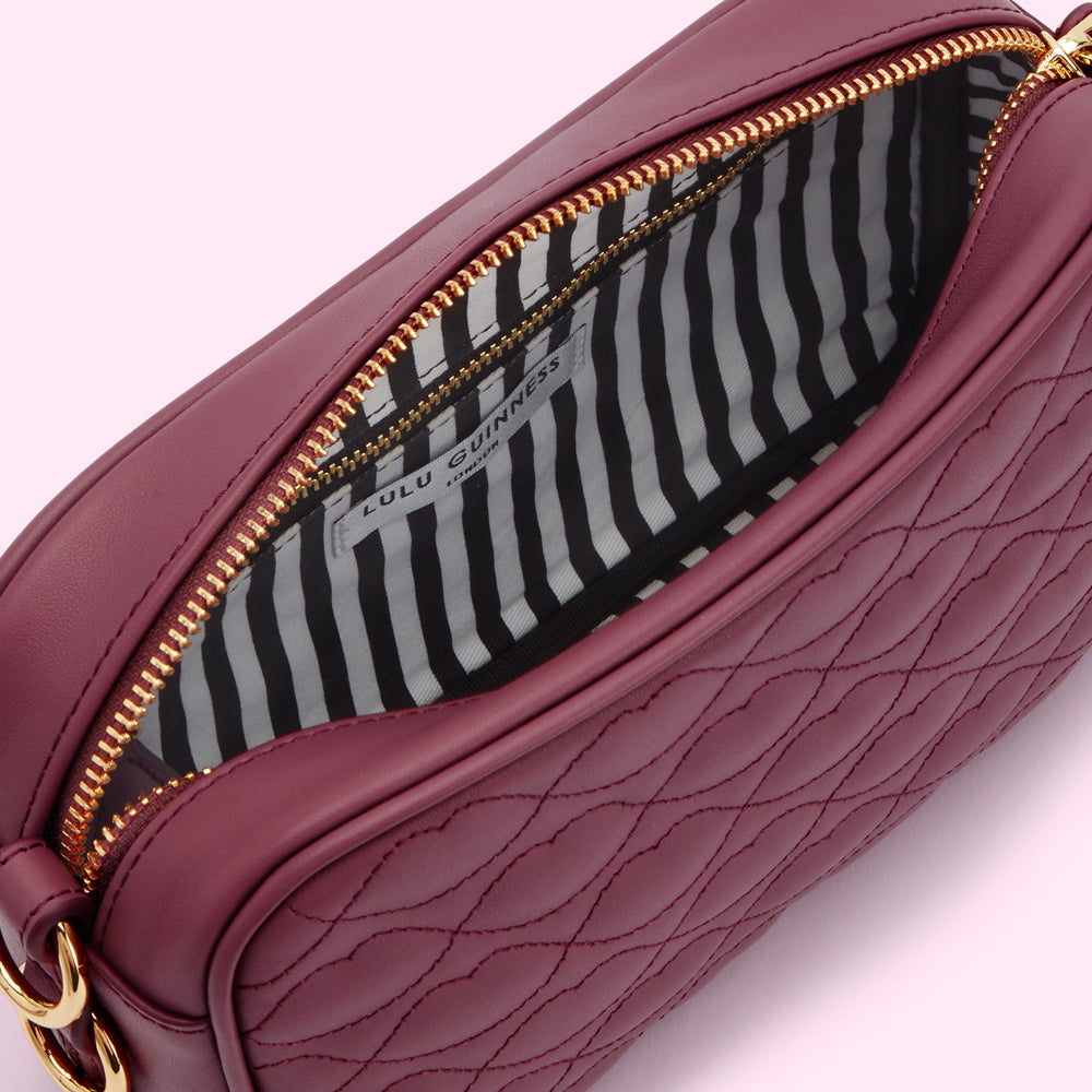 Inside of a burgundy Lulu Guinness quilted crossbody bag, showing the black and white striped lining and gold zipper.
