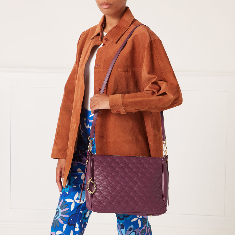 A woman in a brown suede jacket and blue patterned pants carries a quilted purple crossbody bag with a gold charm.
