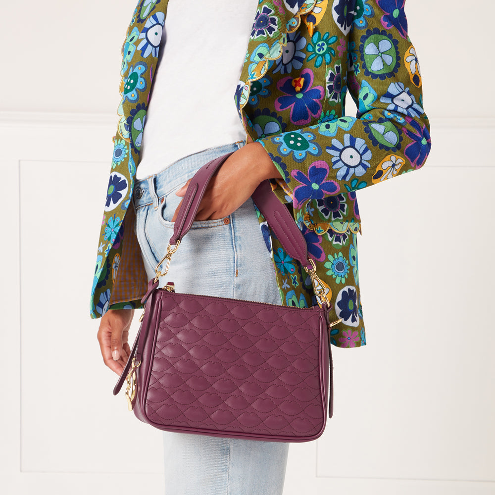 A woman in a floral blazer holds a quilted purple crossbody bag.
