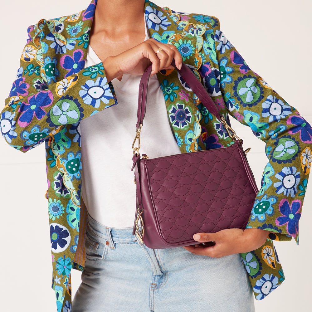 A woman in a floral blazer holds a quilted purple crossbody bag against a white background.
