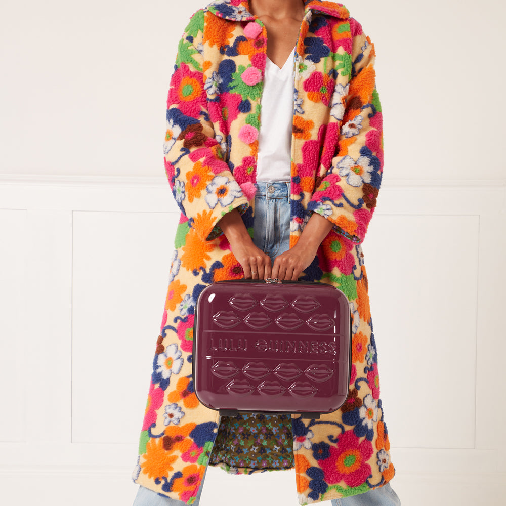 Woman in floral coat holding a burgundy Lulu Guinness Lips suitcase against a white background.
