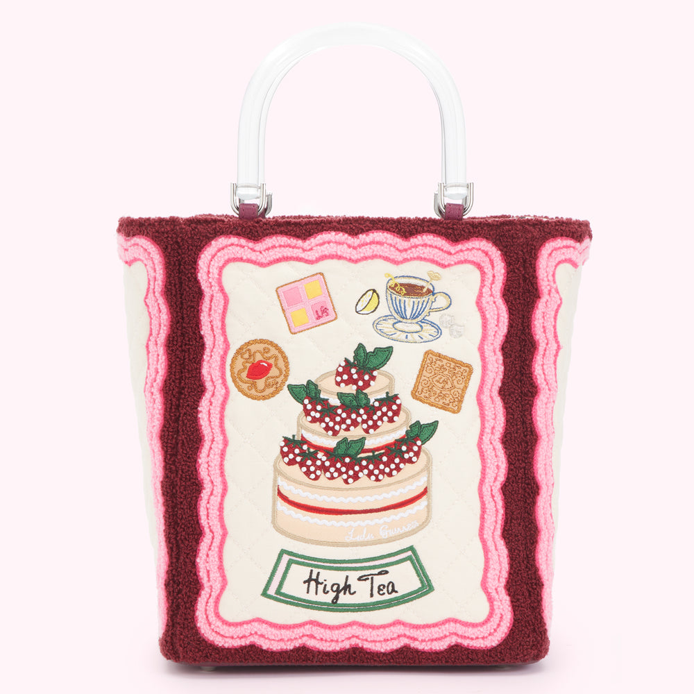 Off-white tote bag with a clear handle, embroidered with a strawberry cake and other high tea treats framed by pink and burgundy scalloped trim. The text "High Tea" is also embroidered on the bag.
