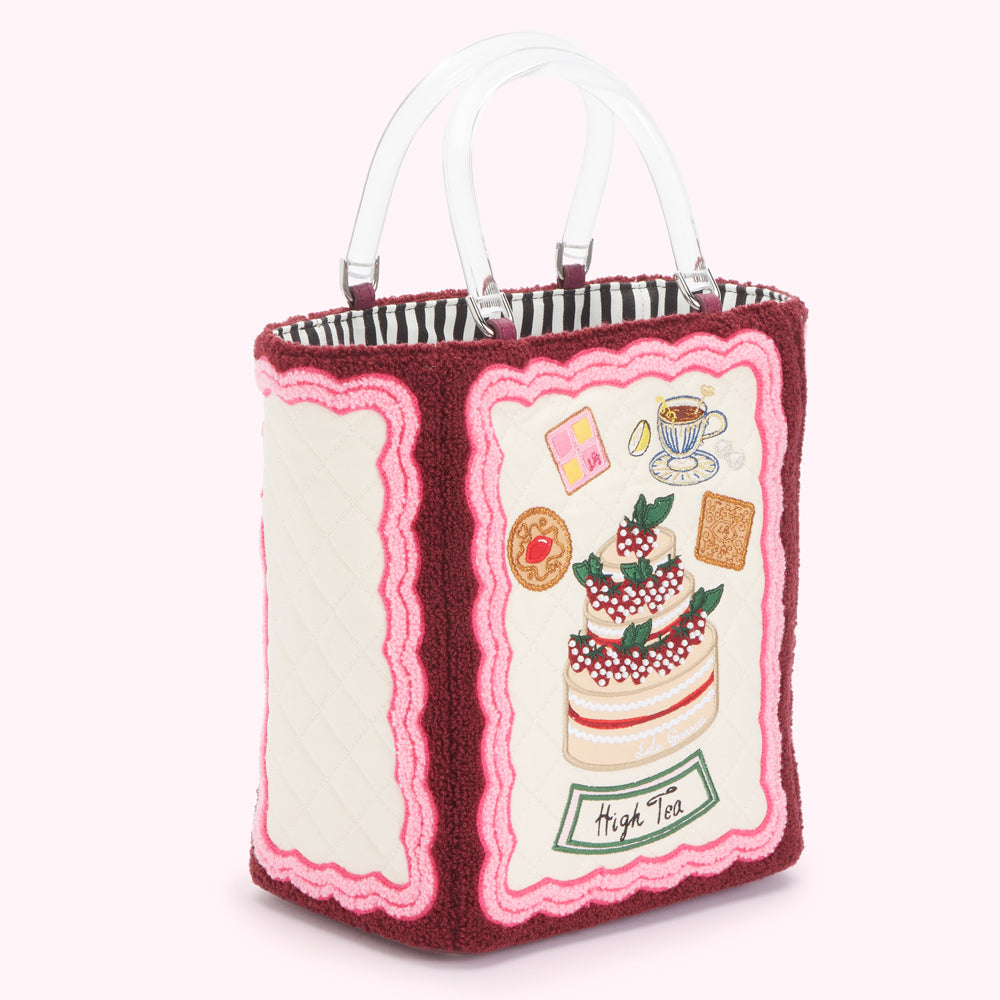 Cream-colored tote bag with maroon and pink trim, featuring embroidered images of pastries, tea, and a strawberry cake, with the words "High Tea" embroidered on the front; the bag has clear acrylic handles.
