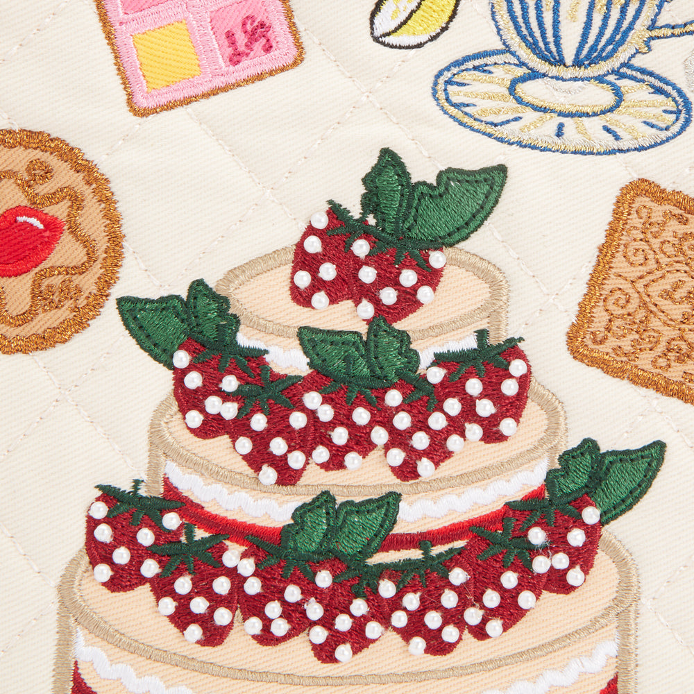 Close-up of embroidered quilted fabric featuring a three-tiered strawberry cake, biscuits, and teacup; off-white background.
