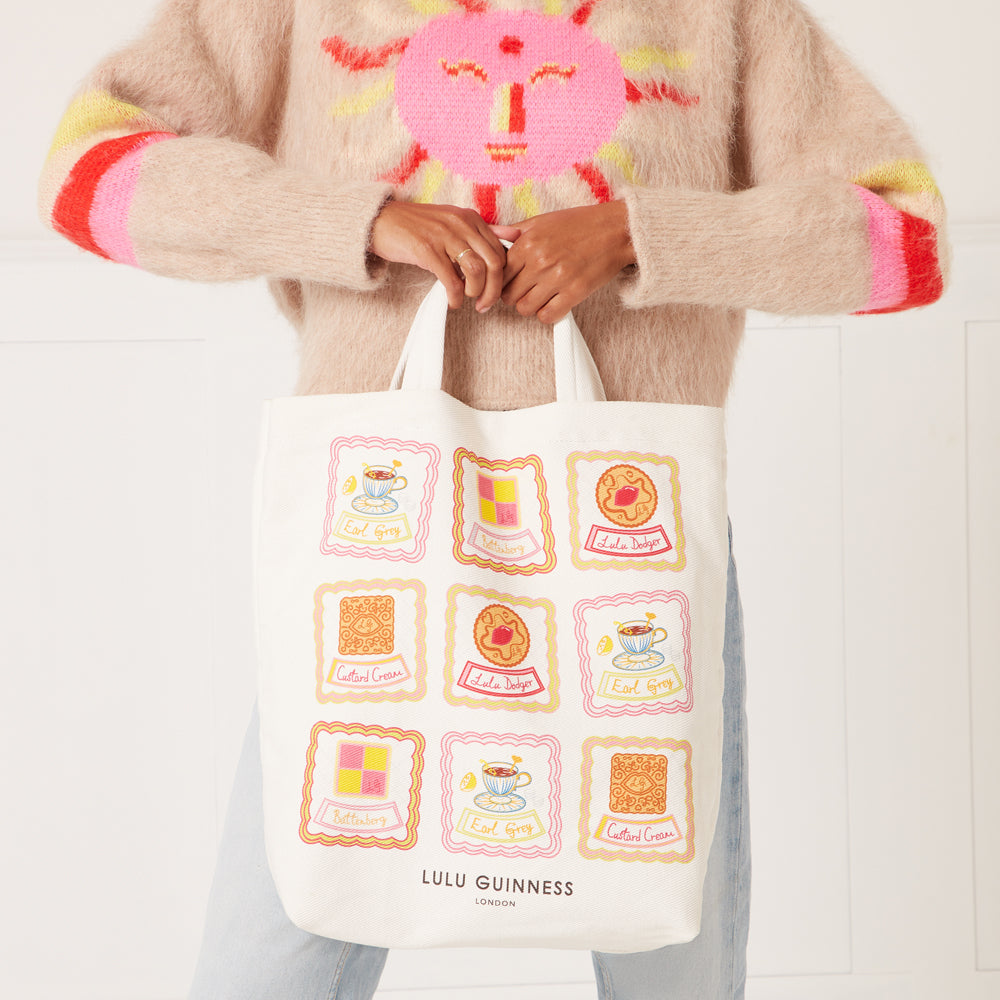 White tote bag with illustrated biscuits and teacups, featuring the Lulu Guinness London logo. A person is holding the bag. The background is off-white.
