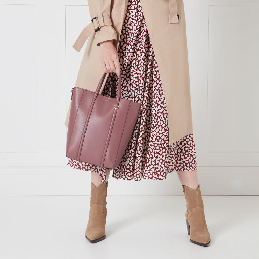 A woman's legs wearing a burgundy floral midi dress, a beige trench coat, and tan ankle boots, holding a dusty rose tote bag.
