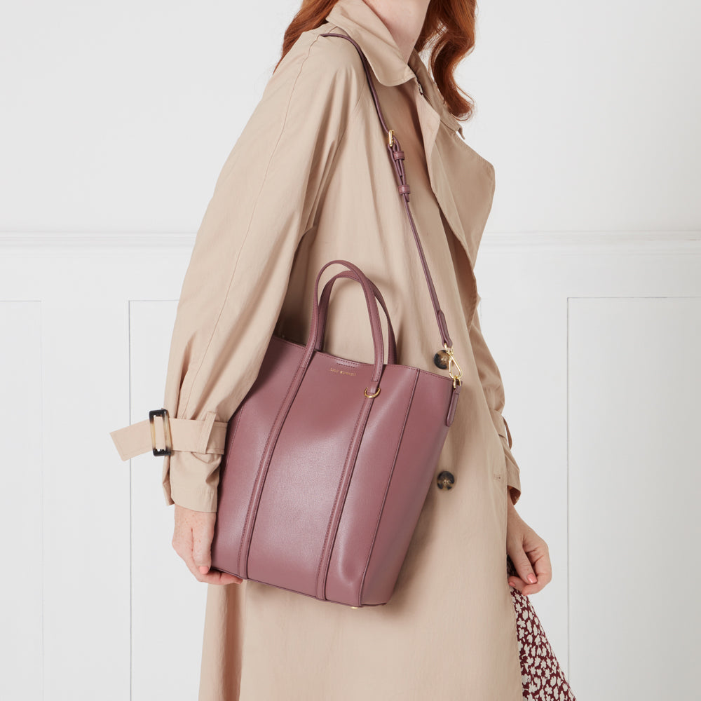 A mauve tote bag with a long strap is held by a woman wearing a beige trench coat.