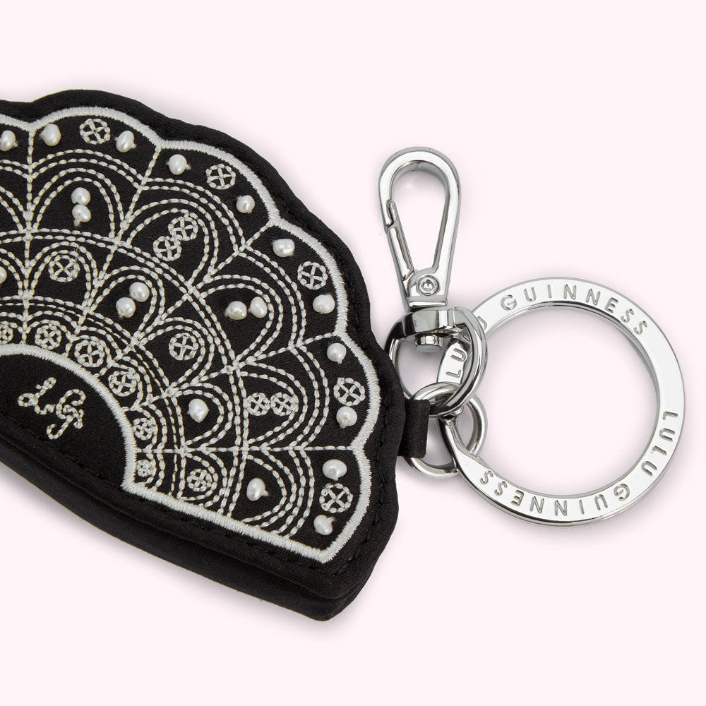 Black leather fan-shaped keyring with white embroidery and faux pearls, attached to a silver keyring with "Lulu Guinness" engraved on it.