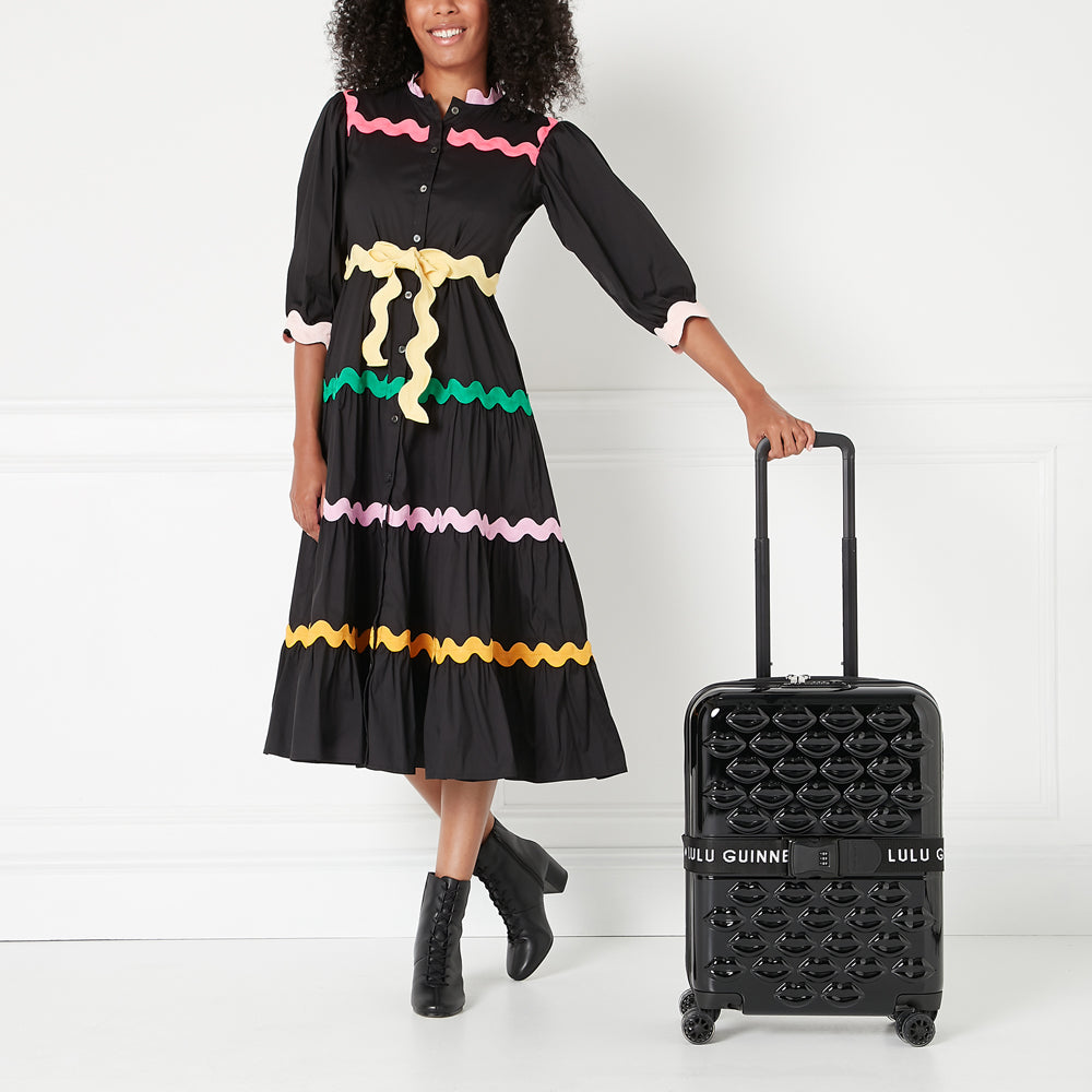 A woman in a black dress with colourful trim stands next to a black carry-on suitcase covered in lip prints. The suitcase has a Lulu Guinness label.