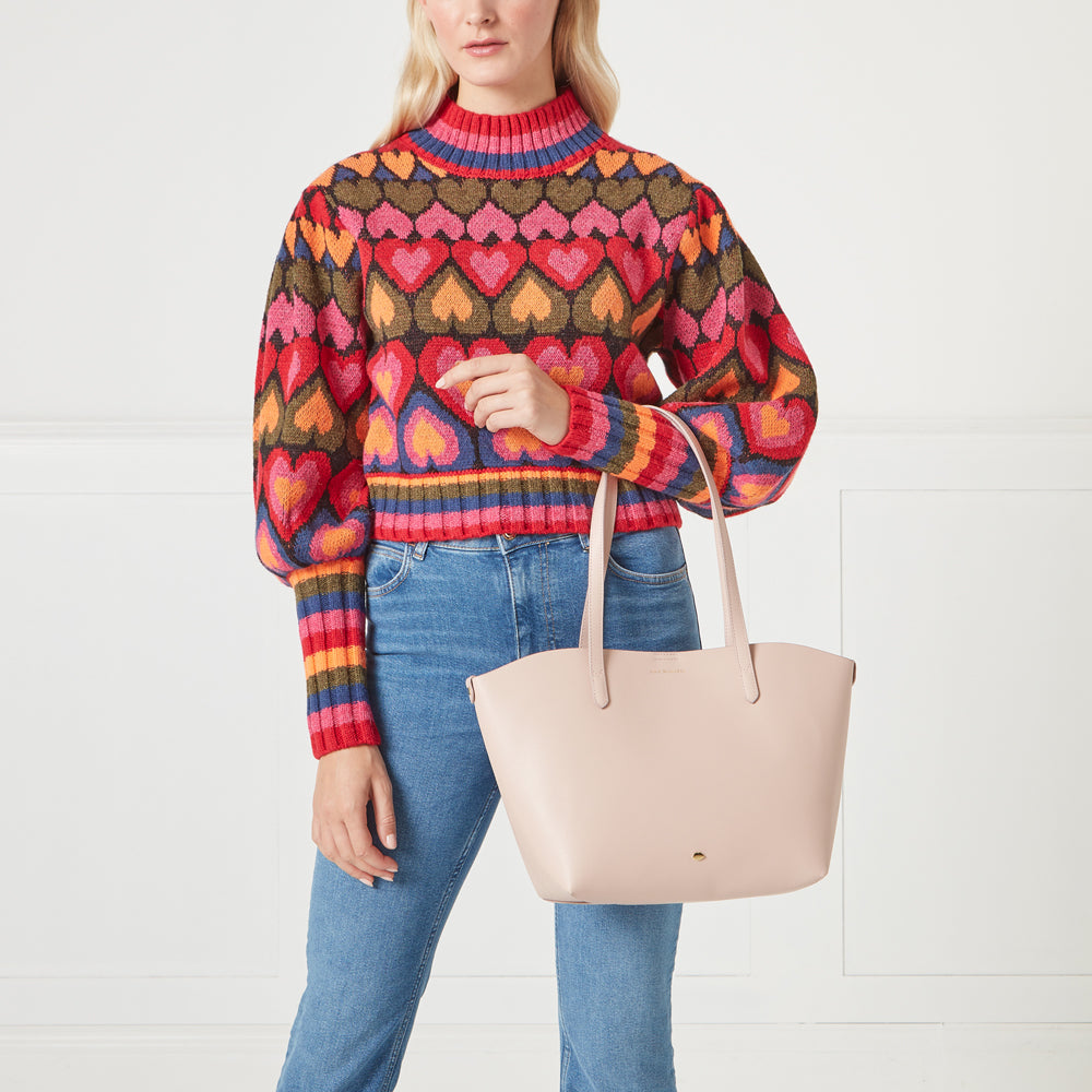 A woman in blue jeans and a multi-coloured heart-patterned sweater holds a blush-pink tote bag.
