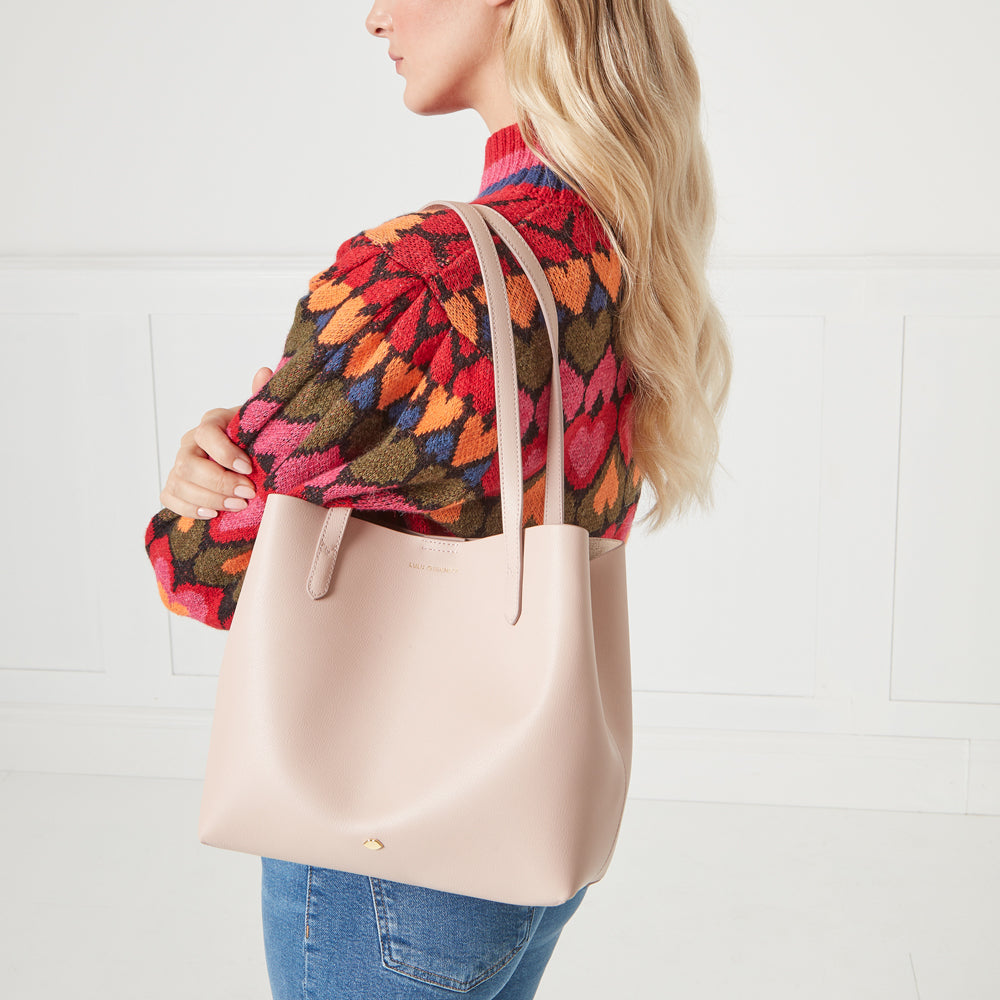 A woman with blonde hair wearing a colourful sweater carries a blush-pink leather tote bag.
