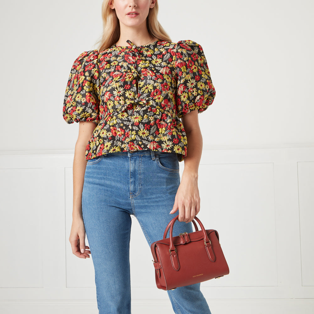 Woman in floral top and jeans holding a dark red handbag against a white background.
