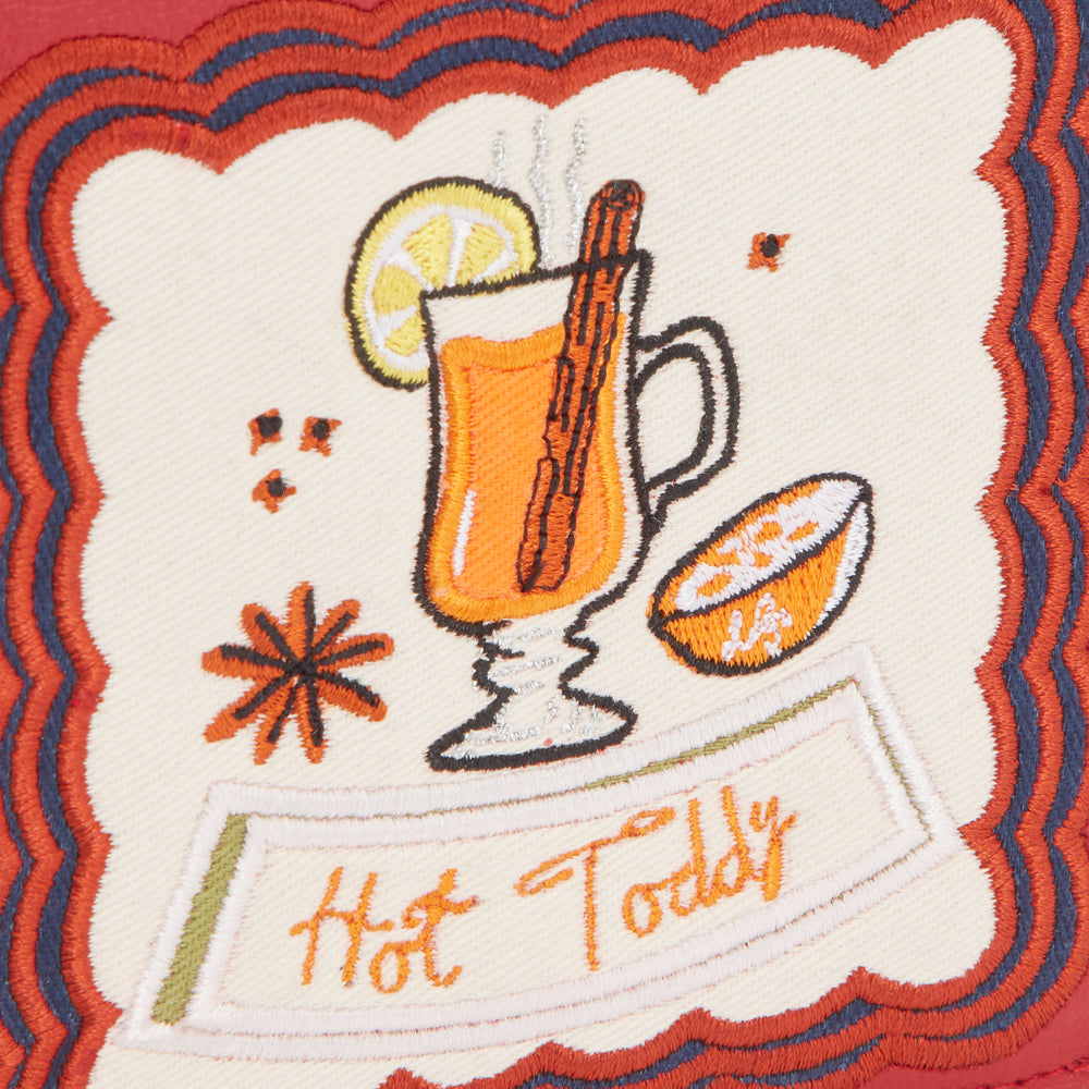Embroidered patch on a red background featuring a whimsical illustration of a hot toddy, complete with orange slice and cinnamon stick; the text "Hot Toddy" is stitched beneath.
