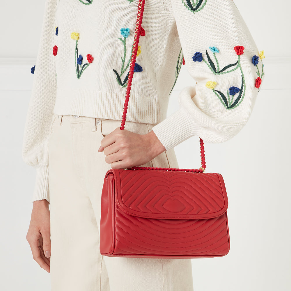 Woman holding a red quilted shoulder bag with gold chain strap, wearing a cream floral embroidered sweater and cream pants against a white background.

