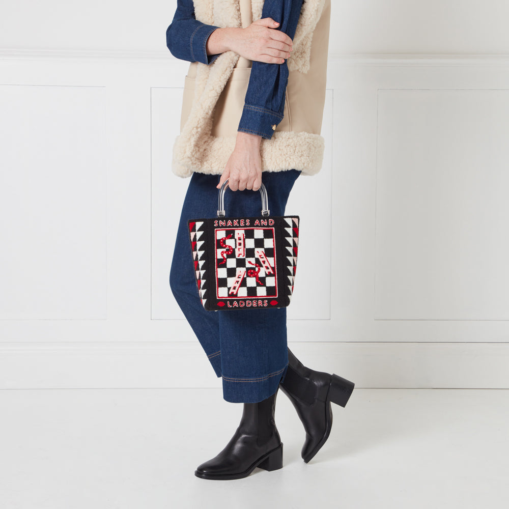 A woman in a denim shirt and cream shearling vest holds a black and white Snakes and Ladders-themed tote bag with a clear acrylic handle.
