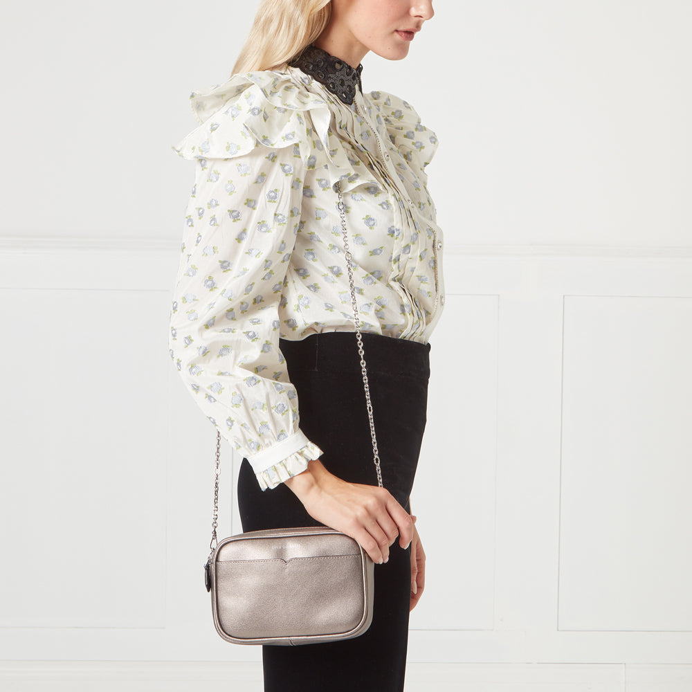 Woman in white floral blouse and black pants holding a metallic silver rectangular crossbody bag against a white background.
