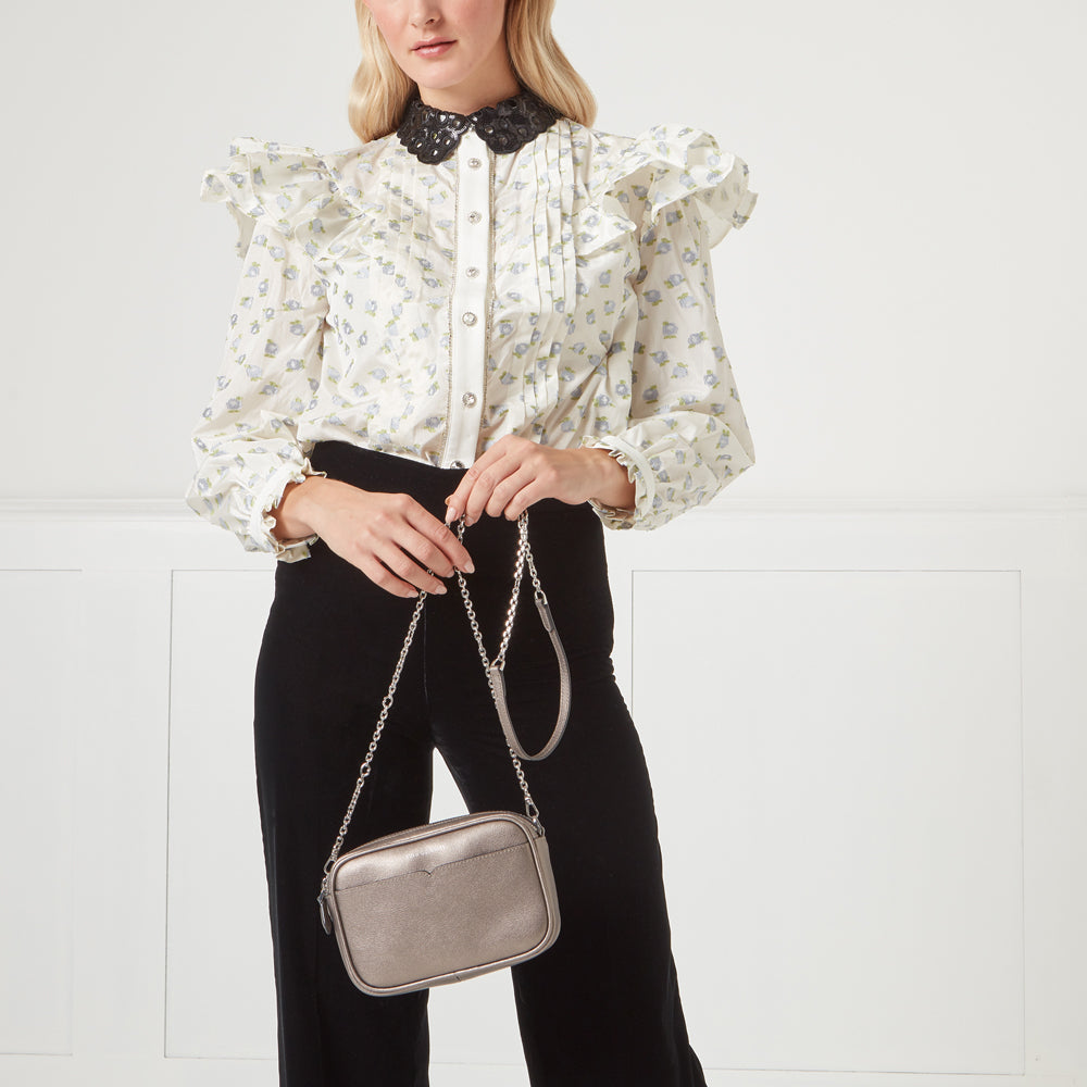 A woman in a floral blouse and black pants holds a metallic silver crossbody bag. The background is white.
