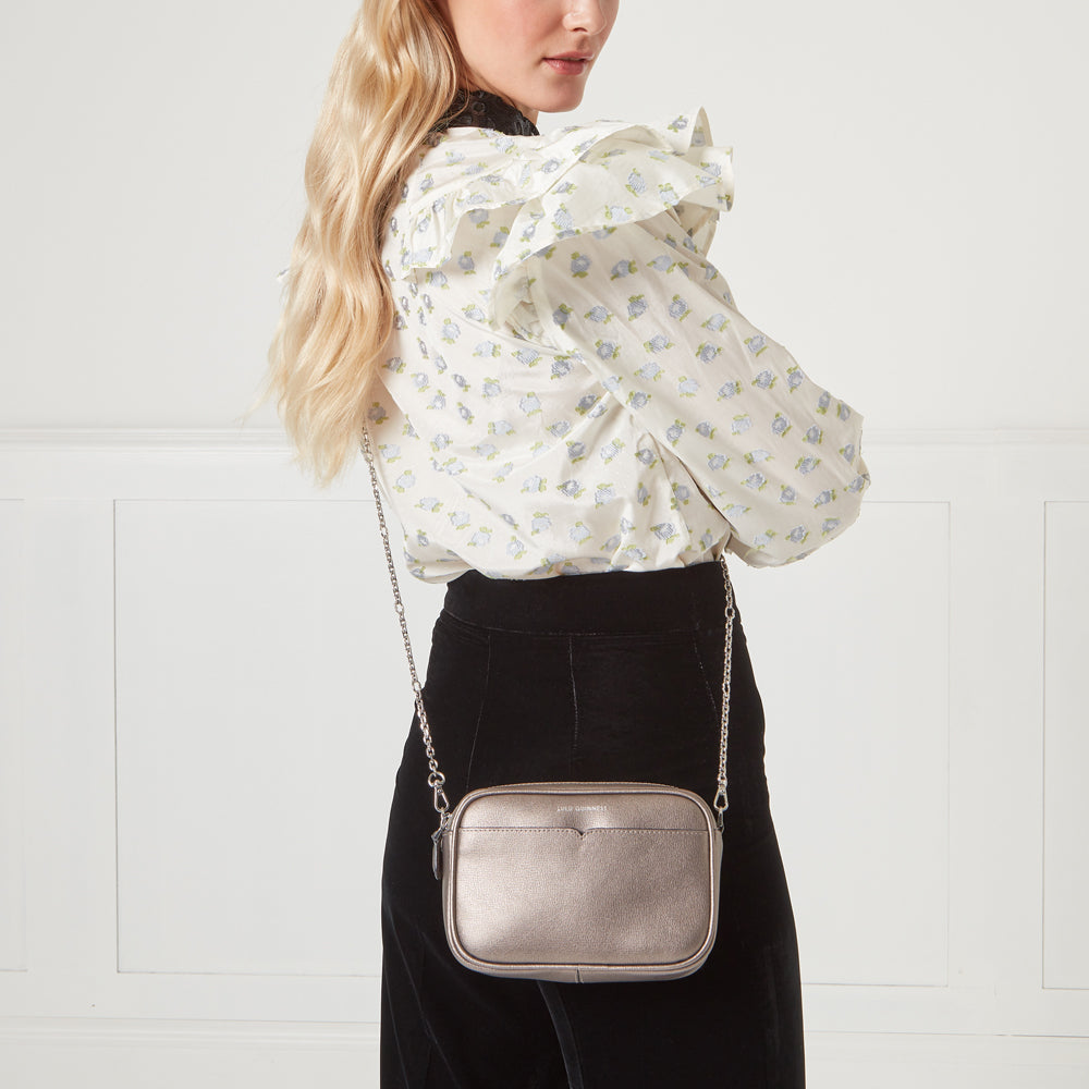 Woman wearing a white floral blouse and black pants with a metallic silver crossbody bag.
