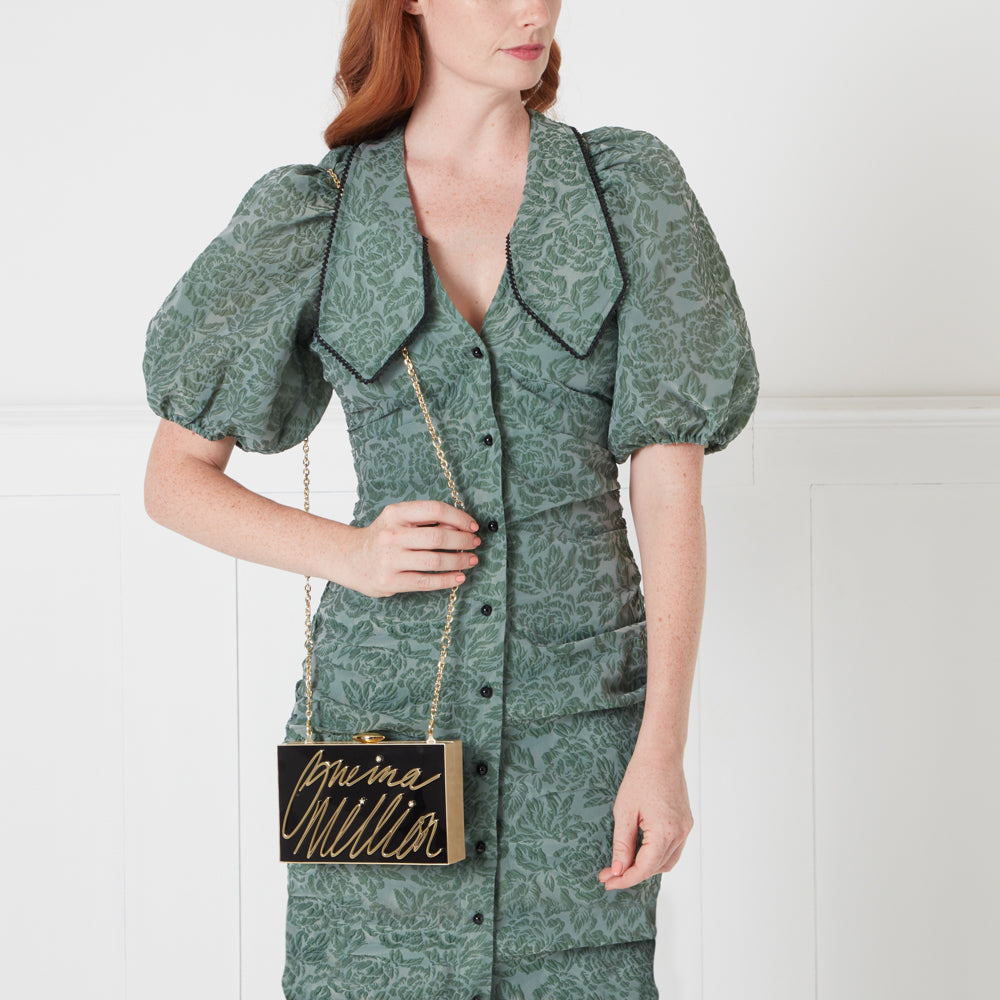 Woman in sage green floral dress holding a black and gold clutch with "One in A Million" written on it.
