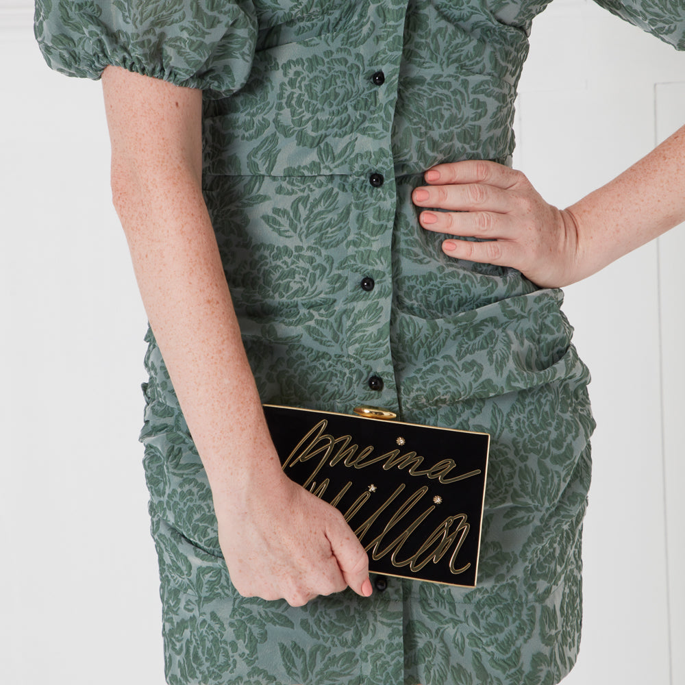 Woman in sage green floral dress holding black clutch that says "One in a Million" in gold lettering.
