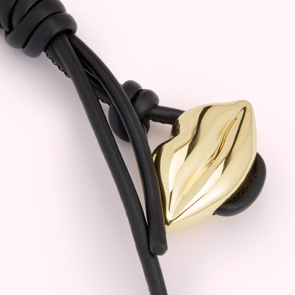 Close-up of a black leather cord bracelet with a gold lip charm.
