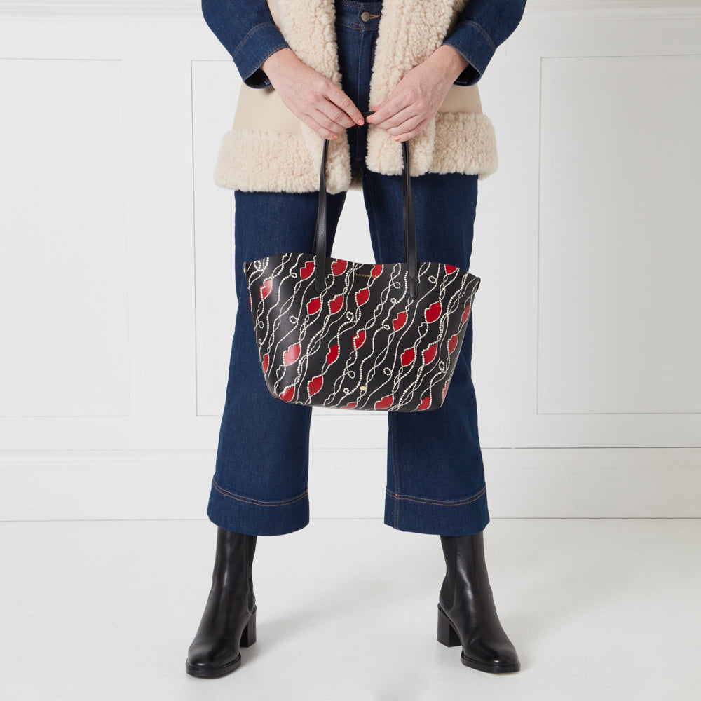 A woman in jeans and a cream-colored shearling vest holds a black tote bag with a red lips pattern.