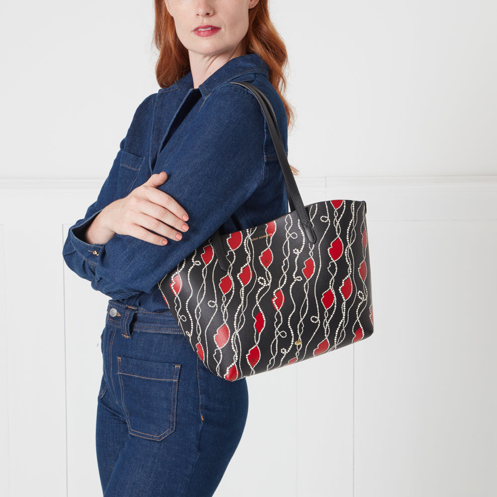 A woman in a denim shirt carries a black tote bag with a red lips and pearl design.
