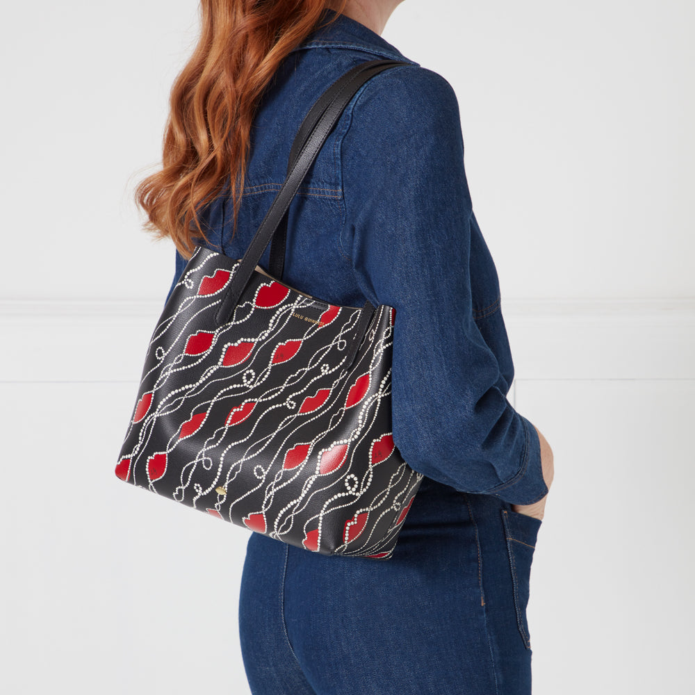 A woman's black tote bag with a red lips and pearl design, worn over her shoulder.

