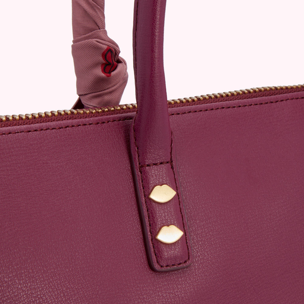 Close-up of a burgundy leather handbag with gold lip-shaped accents on the handle and a pale pink silk scarf detail.
