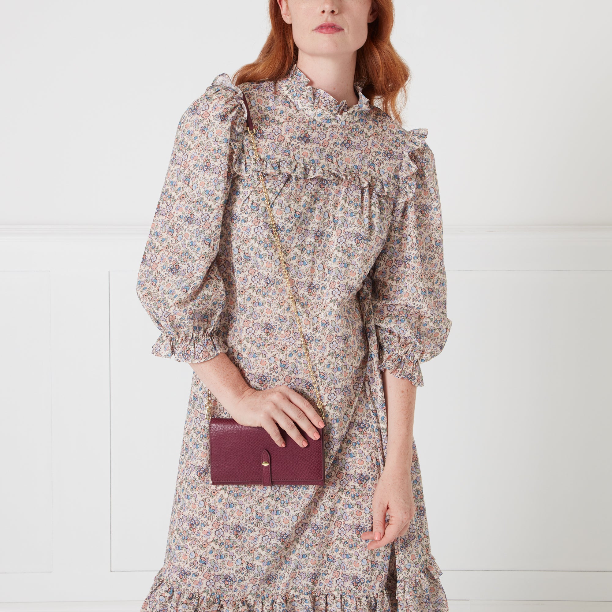 A woman with red hair models a burgundy leather wallet-on-chain against a white background while wearing a floral dress.