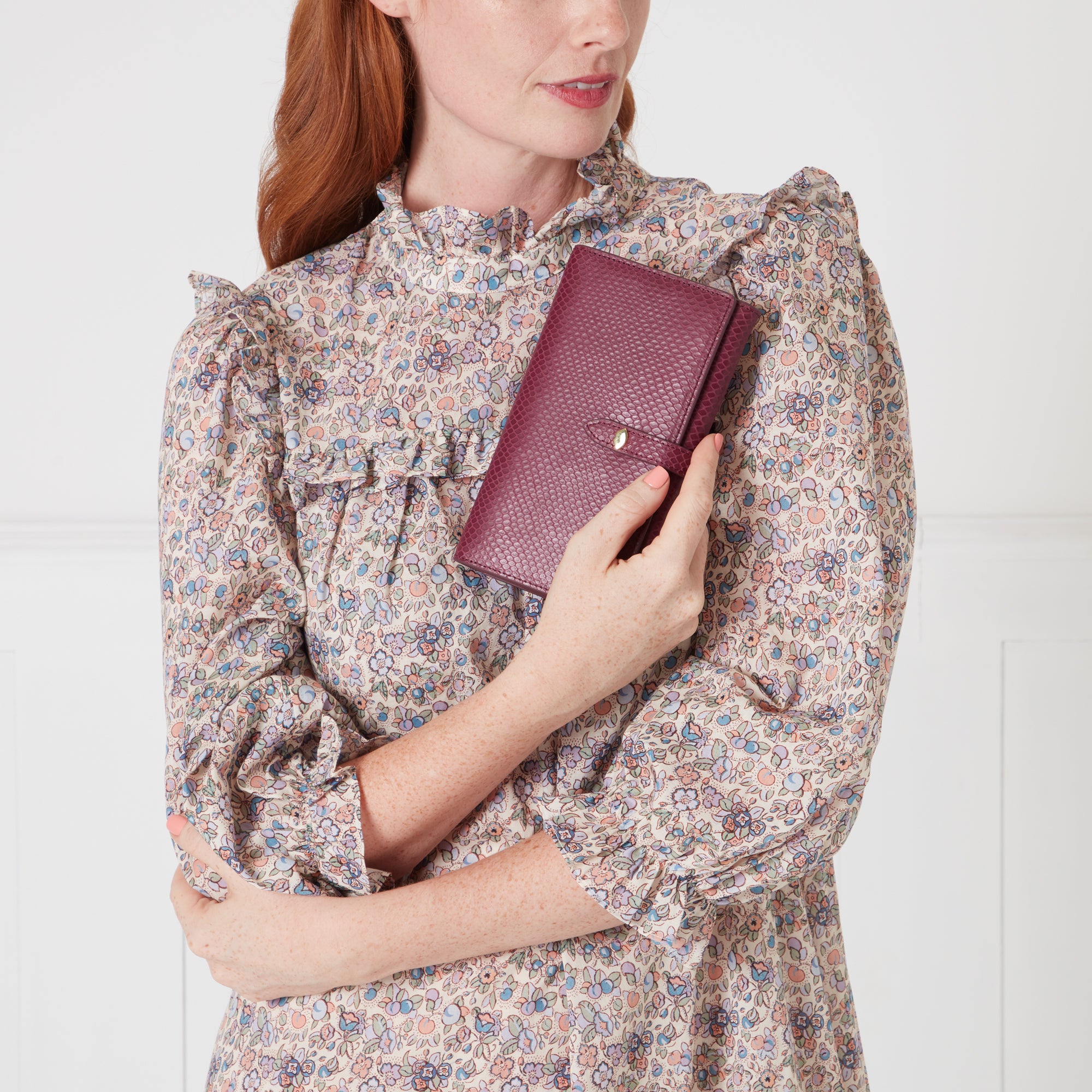 Woman with auburn hair wearing a floral dress holds a burgundy leather wallet. The background is off-white.
