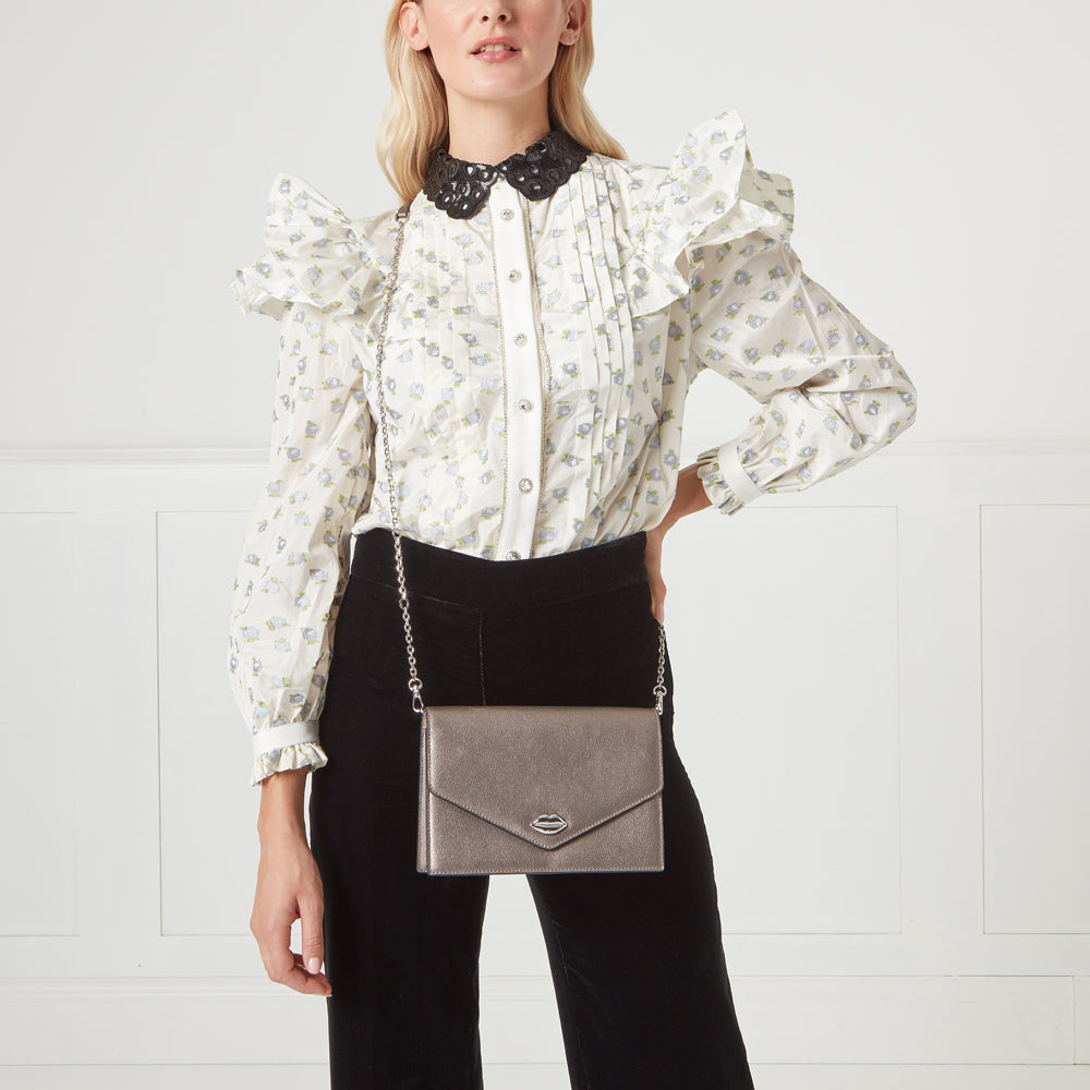 A model wears a white floral blouse and black pants, carrying a metallic silver crossbody bag with a lip-shaped detail. The background is white.
