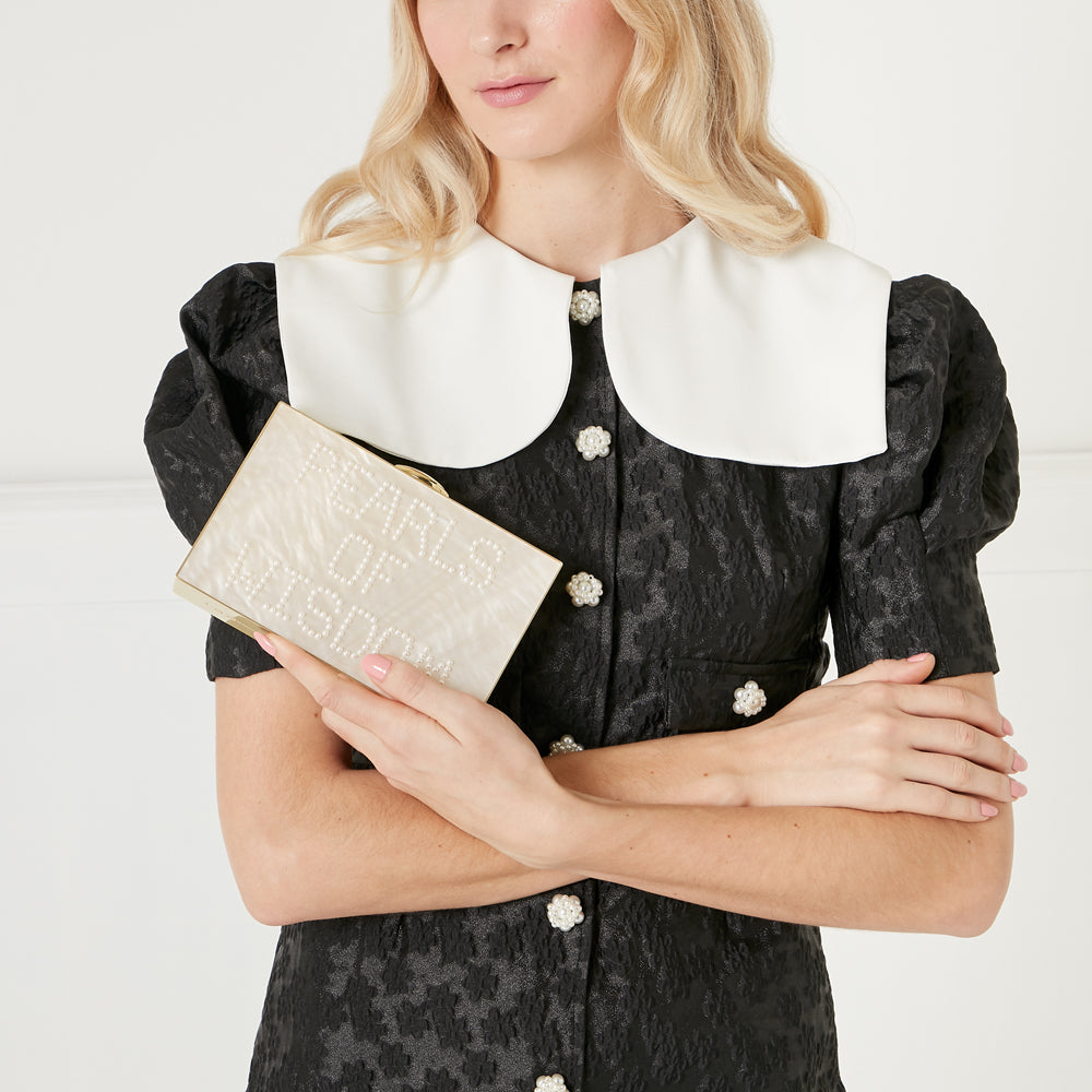 Woman in black dress with Peter Pan collar holding a cream rectangular clutch with "Pearls of Wisdom" written in pearls.
