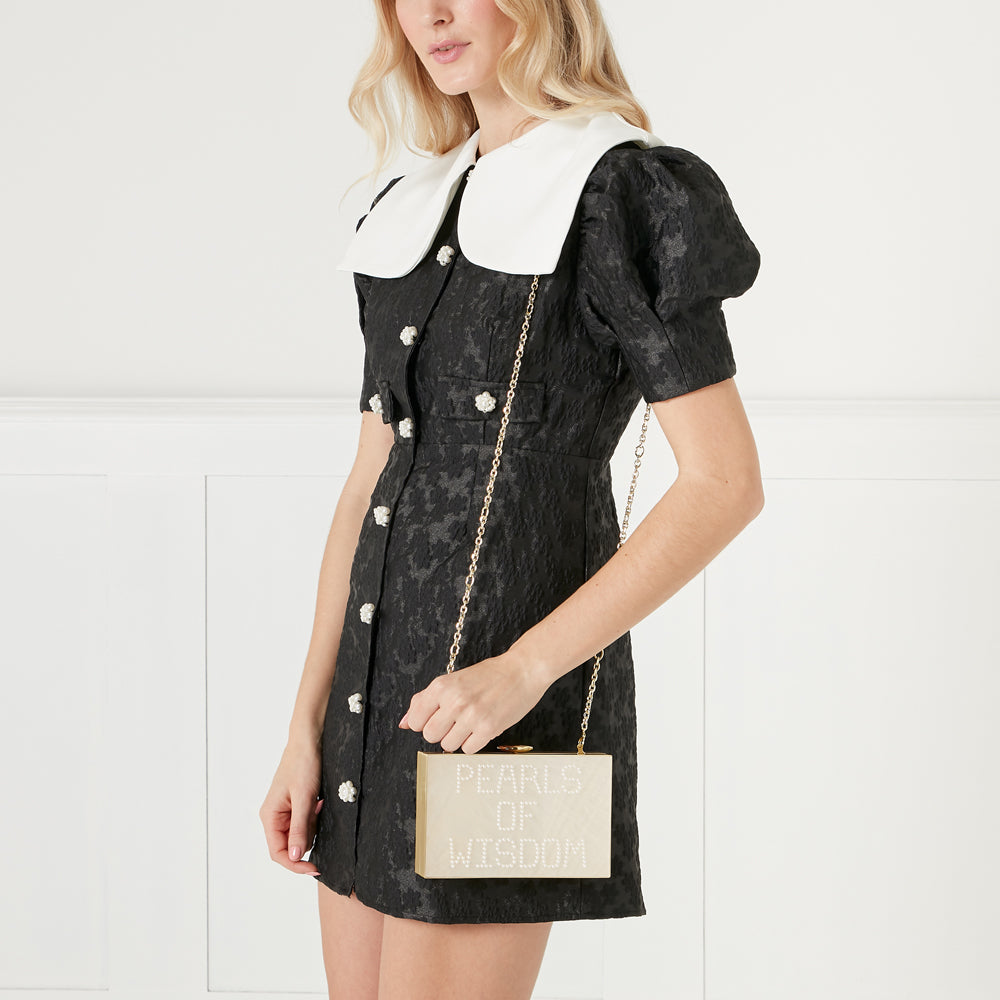 A woman in a black dress with a white Peter Pan collar holds a gold box clutch that says "Pearls of Wisdom" in rhinestones.
