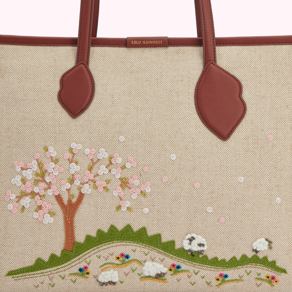 Close-up of a beige Lulu Guinness tote bag with brown leather handles and an embroidered design of a blossoming tree and sheep in a field.