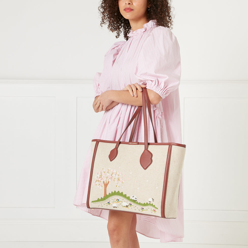 A woman in a pink and white striped dress holds a large beige tote bag with brown leather accents. The bag is embroidered with a whimsical scene of a flowering tree and sheep in a field.