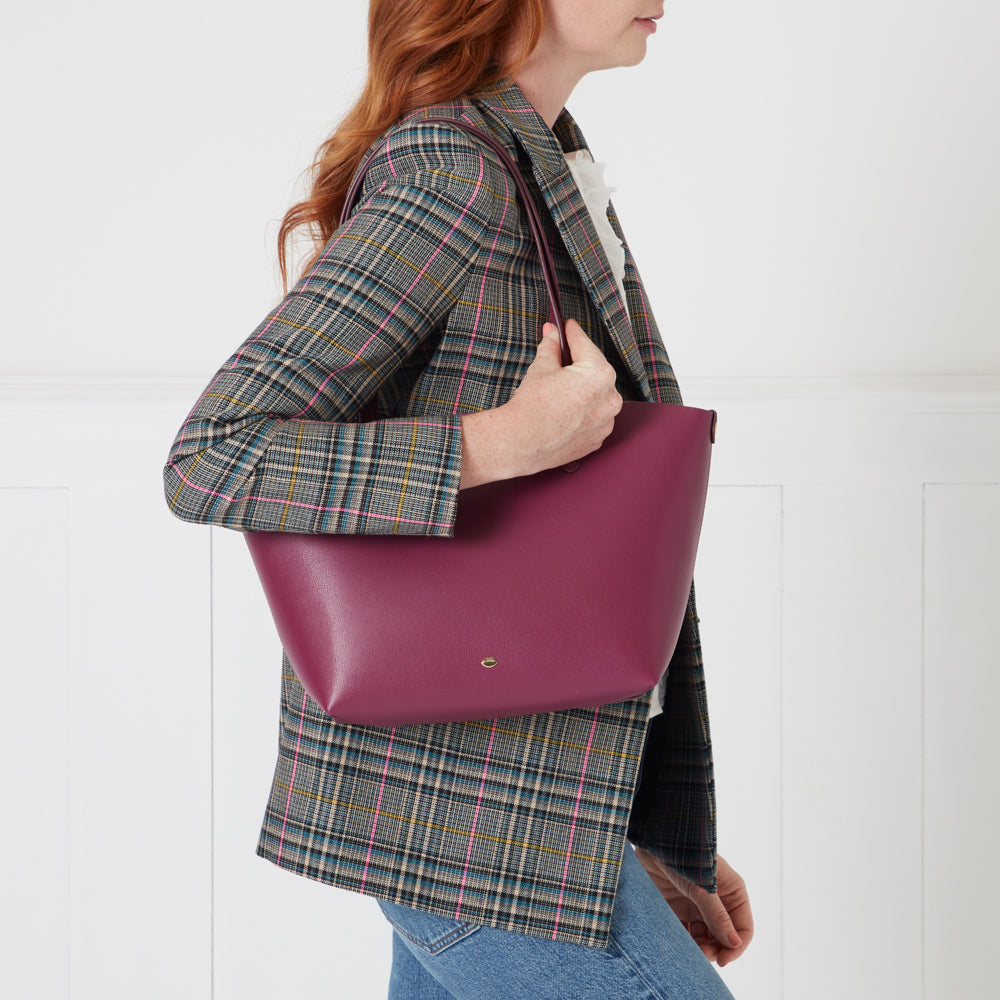 Woman in plaid blazer holding a burgundy tote bag against a white background.
