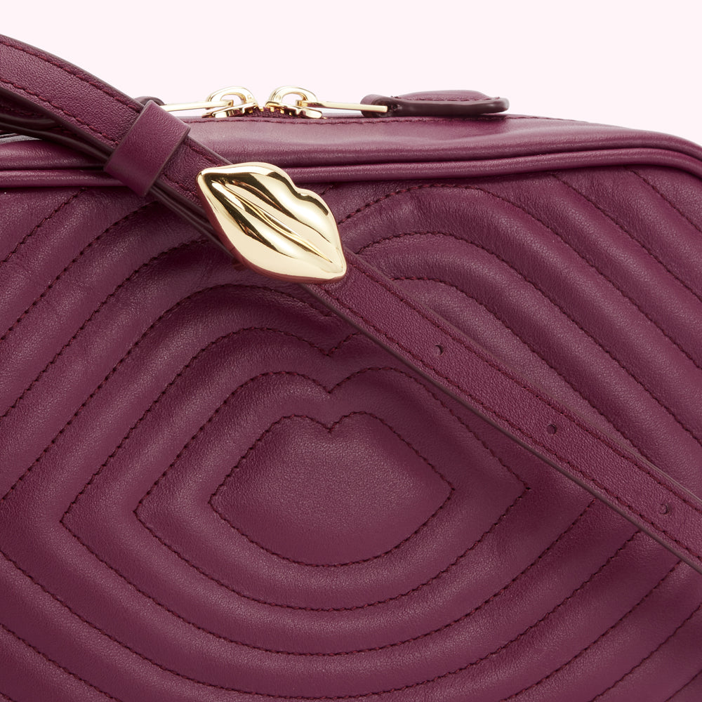 Close-up of a quilted burgundy leather crossbody bag with a gold lip-shaped detail on the strap.
