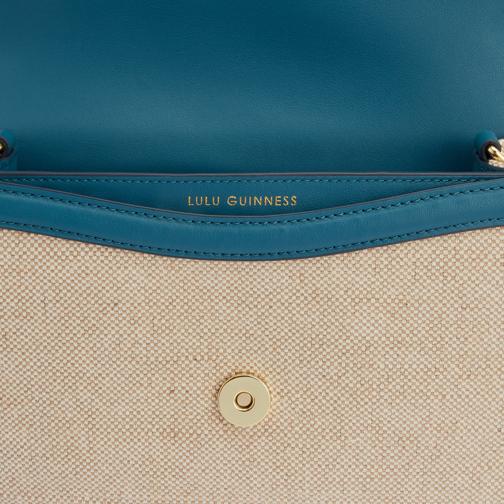 Close-up of the interior of a Lulu Guinness bag, showing beige canvas with teal leather detailing and a gold-toned clasp.
