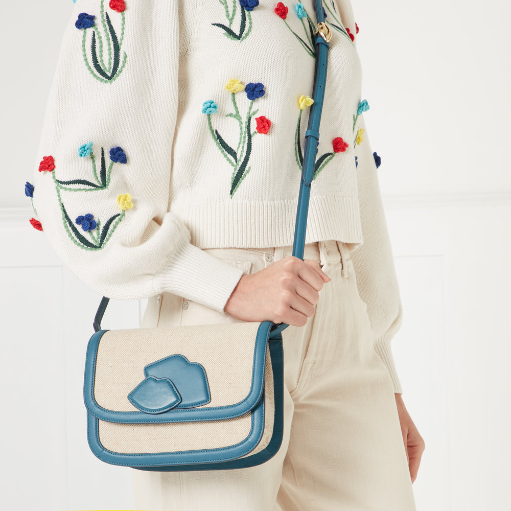 A woman in a cream floral embroidered sweater and cream pants holds a teal and beige crossbody bag.