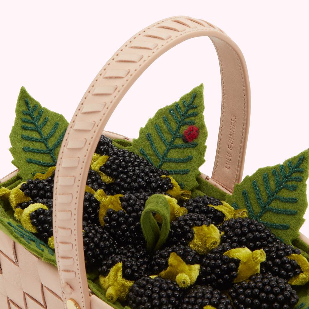 Close-up view of a Lulu Guinness basket bag featuring a pale pink leather handle and a design of black and green embellishments resembling blackberries and leaves.