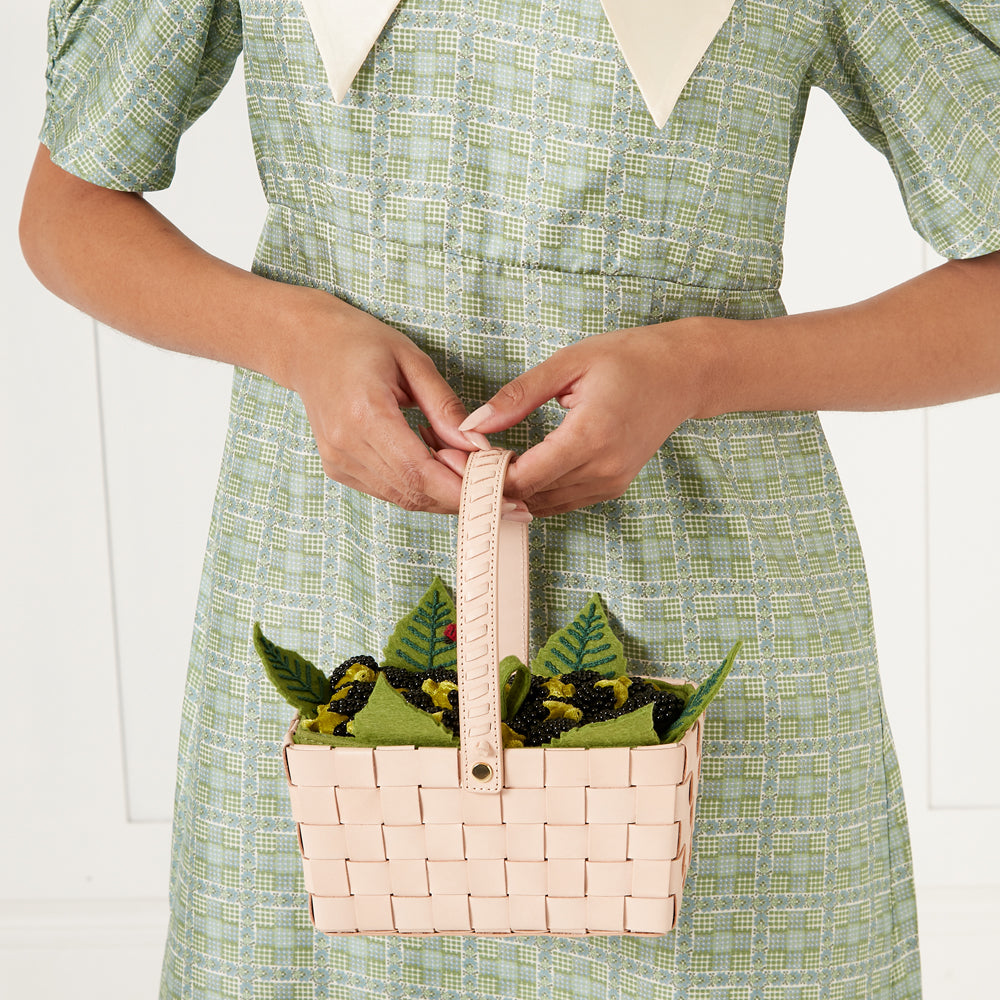 Person in green dress holding a pale pink woven basket decorated with felt fruit and leaves.
