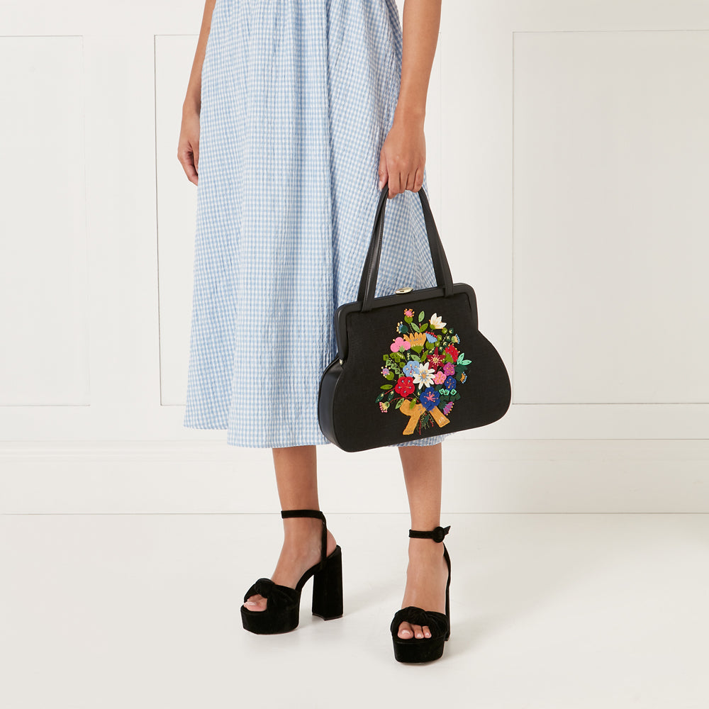 A woman in a light blue gingham dress holds a black handbag embroidered with a colourful floral bouquet. She is wearing black platform heels.