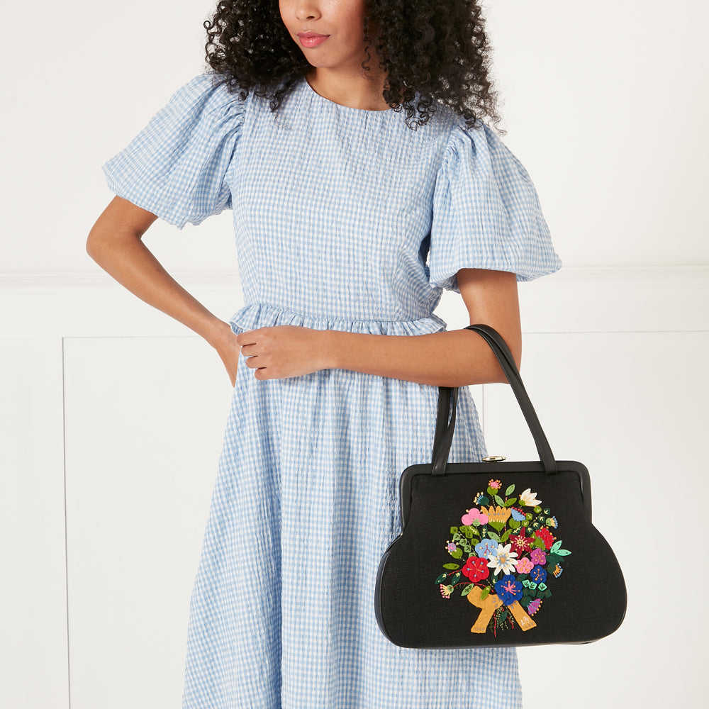 A woman in a light blue gingham dress holds a black handbag embroidered with a colourful bouquet of flowers.