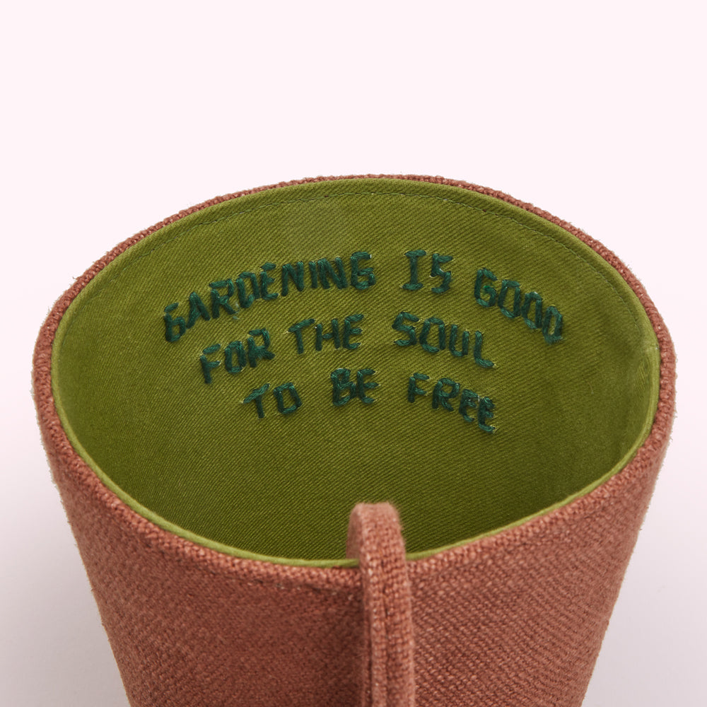 Close-up of a terracotta-coloured plant pot handbag, lined with olive green fabric; the words "GARDENING IS GOOD FOR THE SOUL TO BE FREE" are embroidered in dark green on the inner lining.