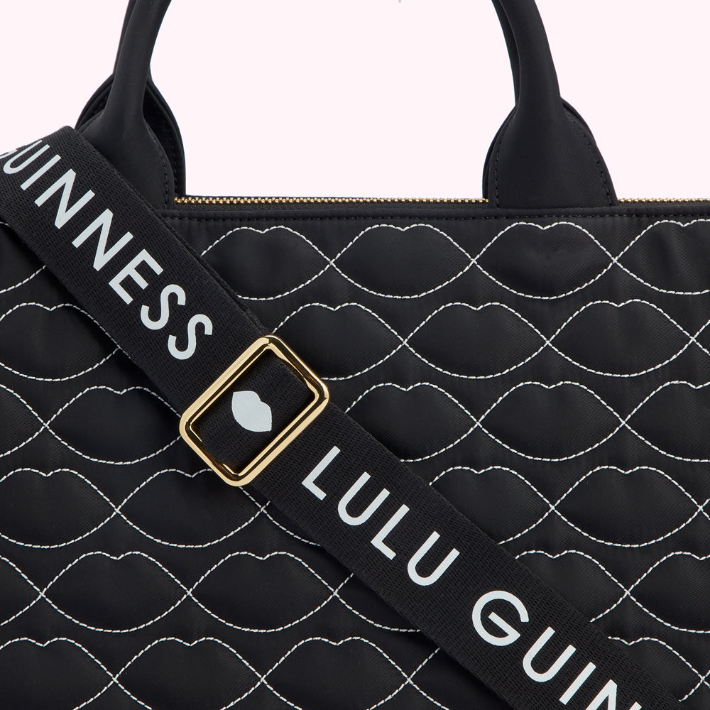 Close-up of a black Lulu Guinness bag with a quilted lip print and a black strap with white "Lulu Guinness" lettering.