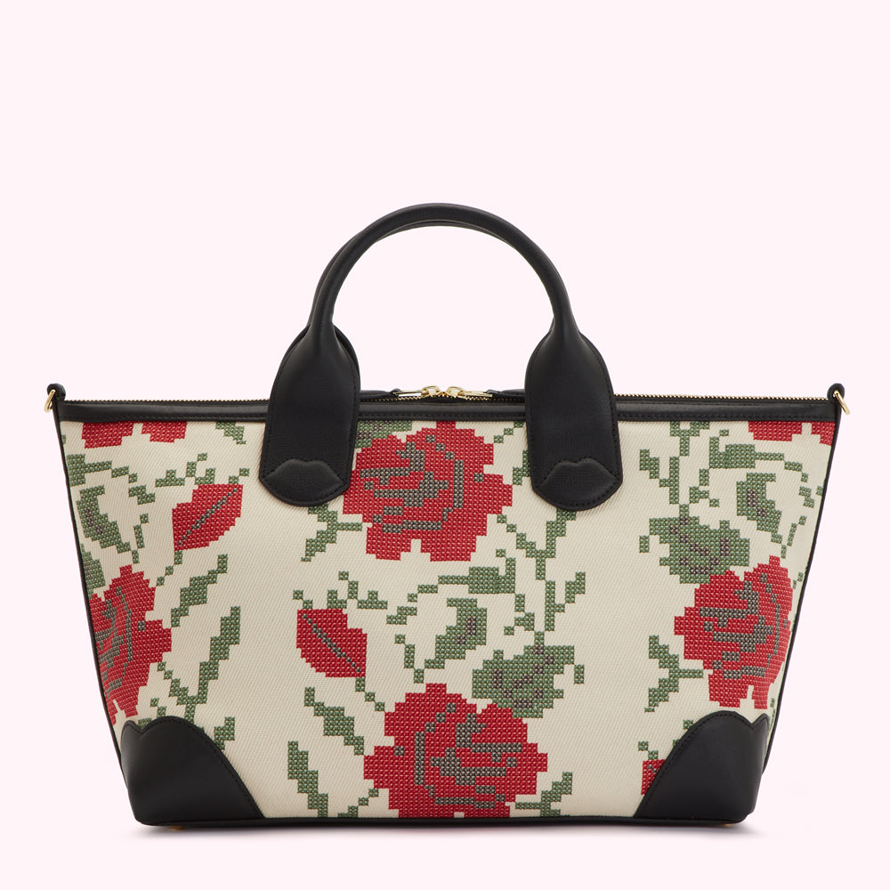Cream-colored handbag with black leather trim and a red and green floral cross-stitch pattern.
