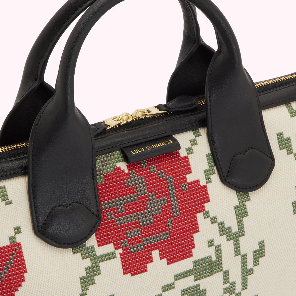 Close-up of a Lulu Guinness handbag with a cream canvas body featuring a red and green cross-stitch floral pattern and black leather handles.
