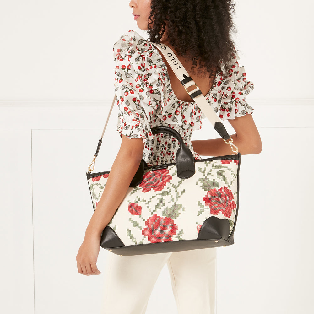 A woman in a floral top carries a large cream-colored tote bag with a red rose pattern and black leather trim. The bag has a beige shoulder strap with the brand name printed on it.