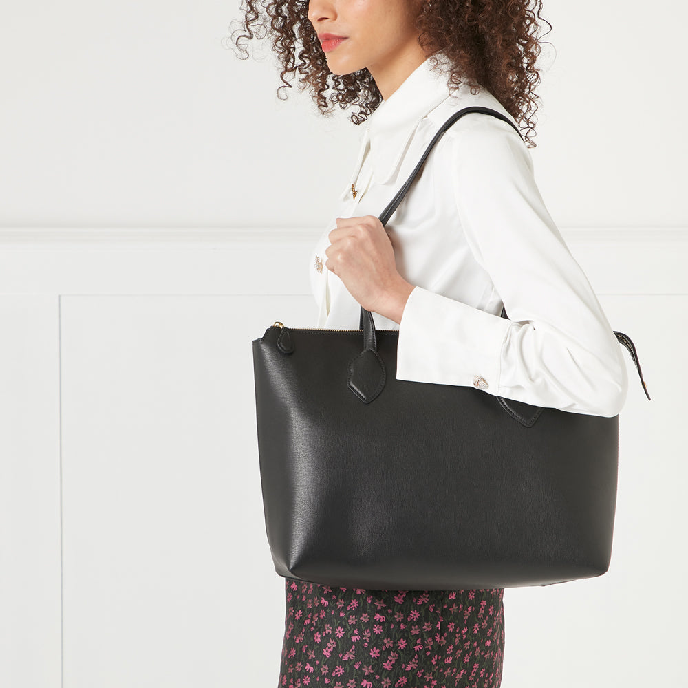 A woman with curly hair holds a black leather tote bag against a white background.
